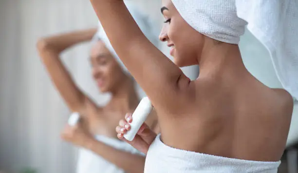 Antiperspirant for dry skin and purity concept. Smiling african american woman in white towel applying deodorant on armpit after shower stands in bathroom in reflection of mirror, panorama, free space