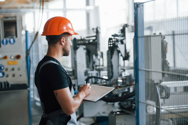 istock With notepad in hands. Industrial worker indoors in factory. Young technician with orange hard hat 1302979190