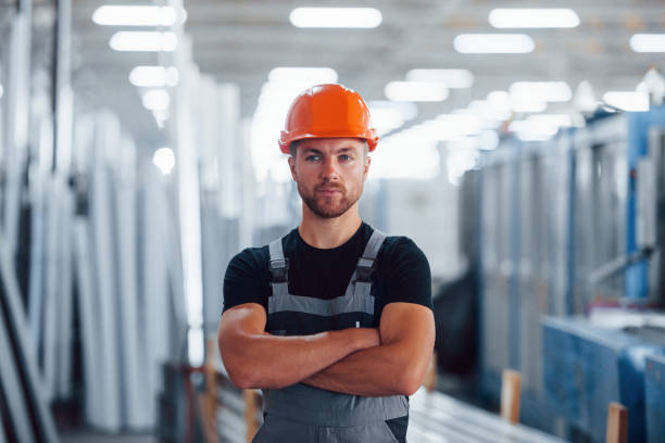 se para con los brazos cruzados. retrato del trabajador industrial masculino en el interior de la fábrica. joven técnico con sombrero duro naranja - manual worker portrait helmet technology fotografías e imágenes de stock