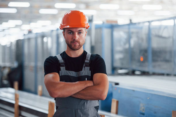 se para con los brazos cruzados. retrato del trabajador industrial masculino en el interior de la fábrica. joven técnico con sombrero duro naranja - manual worker portrait helmet technology fotografías e imágenes de stock