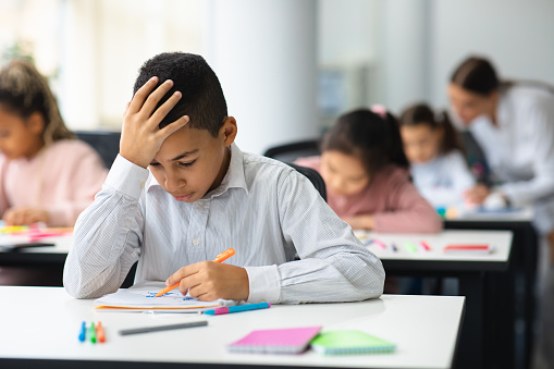 Education And Learning Concept. Portrait of focused small boy sitting at desk in classroom at school, writing in notebook, having test or exam and thinking, touching and grabbing head