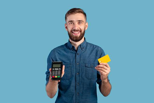 Portrait of happy young guy holding credit card and payment terminal, amiling and looking at camera over blue studio background. Positive millennial man shopping contactless during covid outbreak