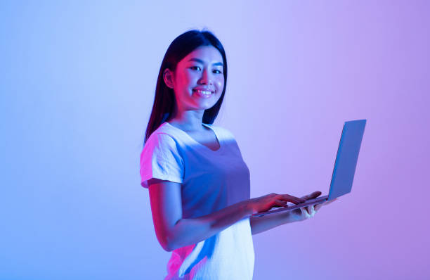 Work remotely online, freelance, study and modern education Work remotely online, freelance, study and modern education. Cheerful confident millennial asian woman looking at camera and typing in laptop for surfing in internet, in neon, studio shot, free space fluorescent photos stock pictures, royalty-free photos & images