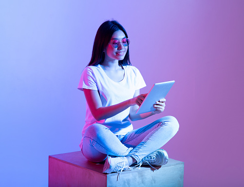 Remote work and study online, application and social networks for chat. Cute busy smiling zoomer asian female in stylish glasses, sits and types in tablet, searching in internet, in neon, studio shot