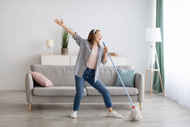 happy woman singing while washing floor at living room, using mop as microphone, having fun during house chores - spring cleaning women cleaning dancing imagens e fotografias de stock