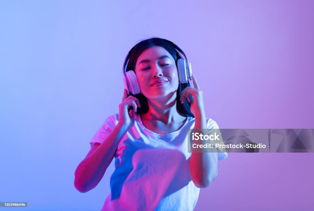 Stay alone at home during covid-19 and relax at spare time with music Stay alone at home during covid-19 and relax at spare time with music. Smiling calm millennial asian lady in modern headphones with closed eyes enjoying song in audio app, in neon, studio shot Headphones Stock Photo