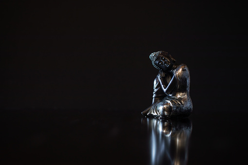 Small black buddha of compassion figurine on dark background with reflection and copy space. For mindfulness and wisdom concepts. Copy space on the right side. Selective focus and added grain. Part of an E+ and S+ series.