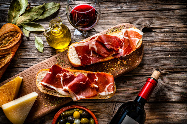 Spanish food: Iberico ham sandwich, Spanish bocadillo de jamon Iberico and red wine Spanish food: overhead view of two bread slices with Iberico ham also called "Bocadillo de Jamón Iberico" with red wine shot on rustic wooden table. Cheeses, olives and pickles and olive oil complete the composition. High resolution 42Mp studio digital capture taken with SONY A7rII and Zeiss Batis 40mm F2.0 CF lens prosciutto stock pictures, royalty-free photos & images