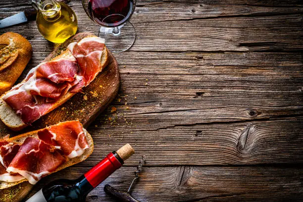 Mediterranean food: overhead view of two Iberico ham sandwich, also called "Bocadillo de Jamón Iberico" on a cutting board shot on rustic wooden table with a red wineglass and a wine bottle. An olive oil bottle and a clay bowl with olives and pickles complete the composition. The composition is at the left of an horizontal frame leaving useful copy space for text and/or logo at the right. High resolution 42Mp studio digital capture taken with SONY A7rII and Zeiss Batis 40mm F2.0 CF lens