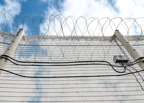 High security prison fence with razor barbed wire on top looking up from ground at solid metal alarmed mesh fencing cables and blue summer sky freedom outside