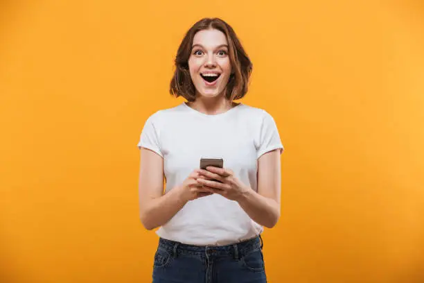 Image of excited young woman standing isolated over yellow background chatting by mobile phone. Looking camera.