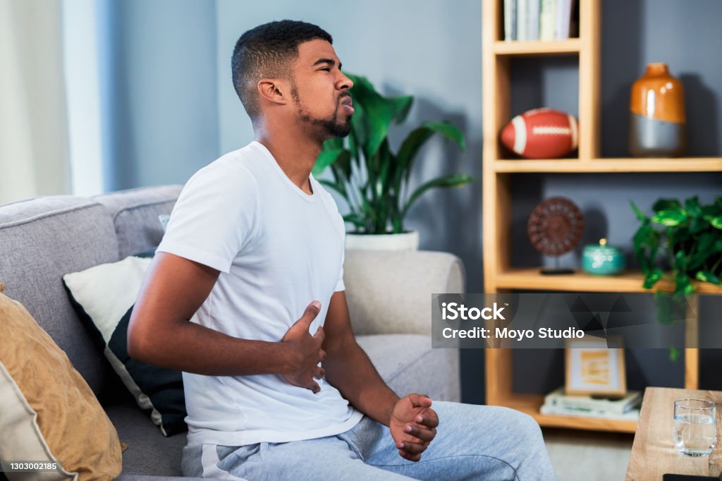 Is there a way to stop this pain? Shot of a young man suffering from stomach pain while lying sitting a sofa at home Stomachache Stock Photo