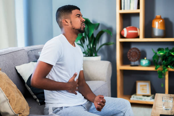 Is there a way to stop this pain? Shot of a young man suffering from stomach pain while lying sitting a sofa at home irritable bowel syndrome photos stock pictures, royalty-free photos & images