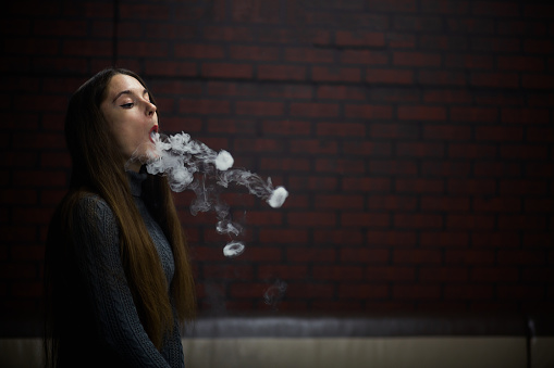 Vaping teenager. Young pretty white caucasian teenage girl with long hair smoking an electronic cigarette indoors. Deadly bad habit. Vape activity.