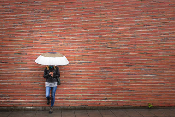 Woman with umbrella is leaning at red brick wall in city. Rain weather on street. Fashionable woman wearing trendy clothes. Copy space