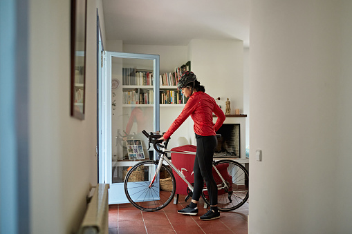 Full length side view of mature Hispanic woman in cycling apparel and helmet wheeling bike outside for daily workout.