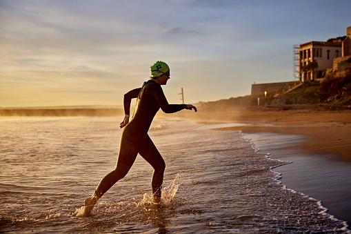 Full length side view of Hispanic sportswoman in wetsuit, swimming cap, and goggles running from water onto beach after workout in Mediterranean.
