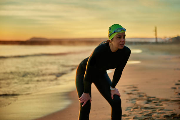Partial front view of Hispanic triathlete in wetsuit, swimming cap, and goggles standing on beach after workout in Mediterranean Sea at sunrise.