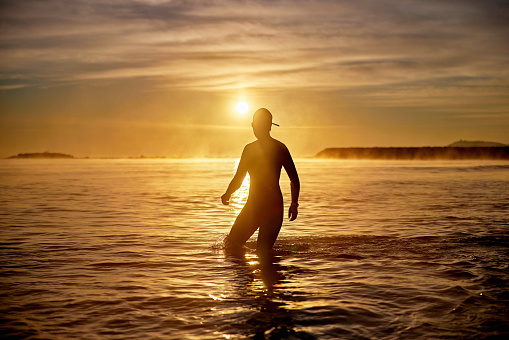Partial view of swimmer in silhouette wearing wetsuit, swimming cap, and goggles as she walks in water following early morning workout.