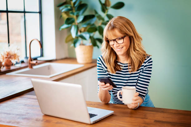 istock Mature woman using her mobile phone and laptop while sitting in her kitchen at home 1303010696