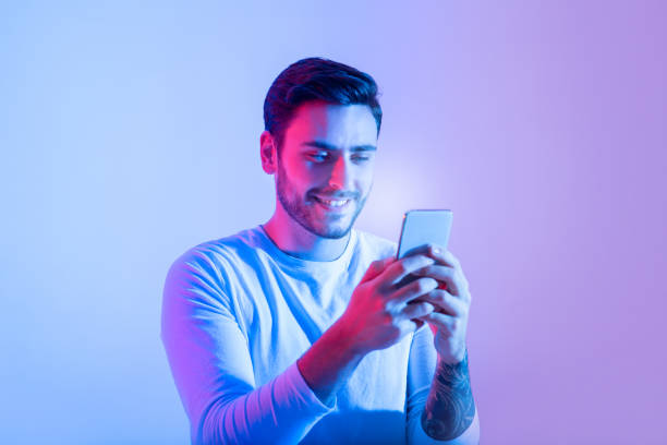 Addiction to game, chat in social networks and online chat Addiction to game, chat in social networks and online chat. Smiling excited young guy with bristle, looks at glowing phone and typing message, surfing in internet in neon, studio shot, free space mobile phone lights up stock pictures, royalty-free photos & images