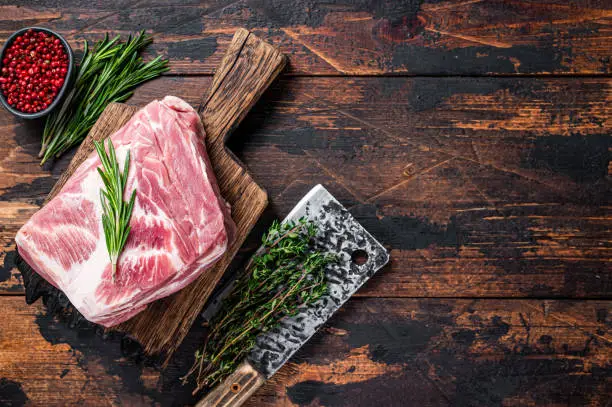 Pork Shoulder raw meat for fresh steaks on wooden cutting board with butcher cleaver. Dark wooden background. Top view. Copy space.