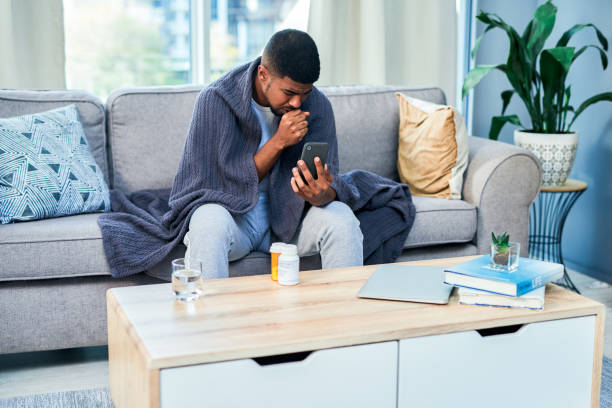 Video calling his doc for a virtual consult Shot of a young man using a cellphone while feeling ill at home cold and flu man stock pictures, royalty-free photos & images