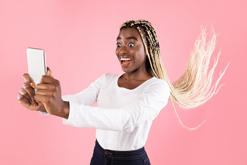 Portrait of excited emotional funny african american woman holding and using mobile phone, taking selfie, blogging, reading surprising news, having her hair fly, copy space, pink studio background