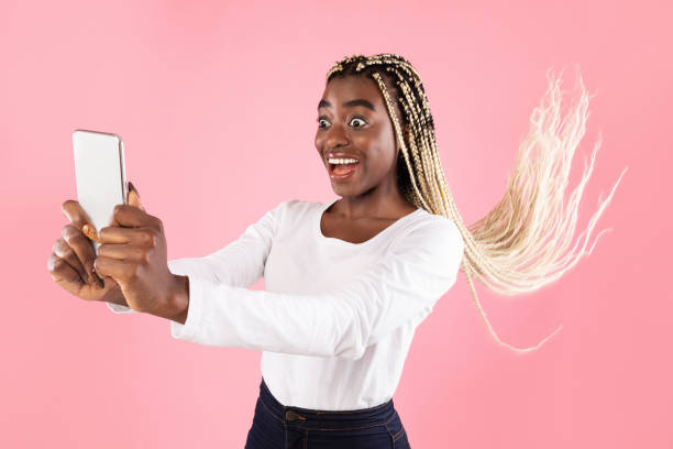 Woman holding smartphone with her long hair flying Portrait of excited emotional funny african american woman holding and using mobile phone, taking selfie, blogging, reading surprising news, having her hair fly, copy space, pink studio background windspeed stock pictures, royalty-free photos & images