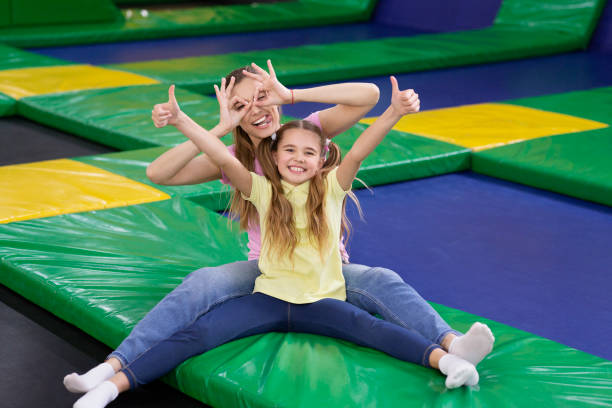 Playful silly mom with her teen daughter sitting at trampoline area of entertainment centre, showing thumbs up gesture Playful silly mom with her teen daughter sitting at trampoline area of entertainment centre, showing thumbs up gesture. Mother and her child recommending to visit indoor kids playground trampoline stock pictures, royalty-free photos & images