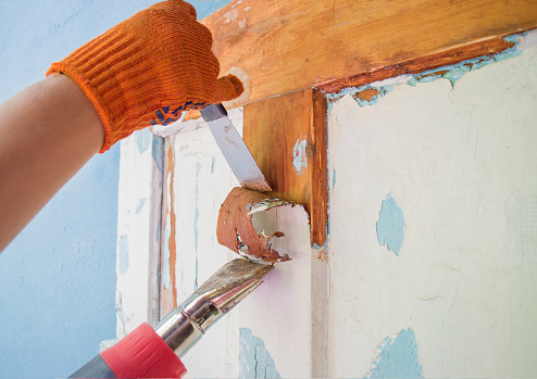 The process of cleaning an old wooden door from paint layers. Removal of the coating with a hair dryer and a spatula. Worker hands in orange gloves with a tool. Household renovation.
