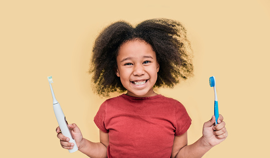 Smiling African American little girl holds manual and electric sonic toothbrushes. Child selects kind of toothbrush, isolated on pastel background