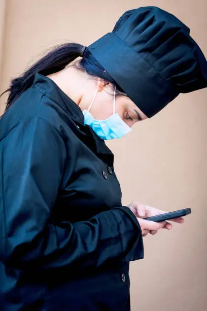 Photo of Crestfallen young female chef looking at her smartphone wearing a protective face mask