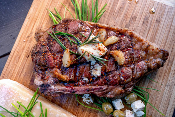 Ribeye Steak Just Off A Charcoal Grill Resting On A Wood Cutting Board, Seasoned With Rosemary, Garlic Cloves, Butter, Cheese, Olives And Paprika Ribeye Steak Just Off A Charcoal Grill Resting On A Wood Cutting Board, Seasoned With Rosemary, Garlic Cloves, Butter, Cheese, Olives, And Paprika grass fed stock pictures, royalty-free photos & images