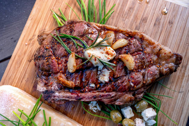 bife de ribeye apenas fora de uma grelha de carvão descansando em uma tábua de corte de madeira, temperado com alecrim, cravo de alho, manteiga, queijo, azeitonas e páprica - rib steak - fotografias e filmes do acervo