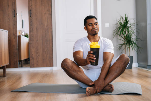istock African-American man sitting cross-legged and relaxing on exercise mat after workout, drinking water 1303043008