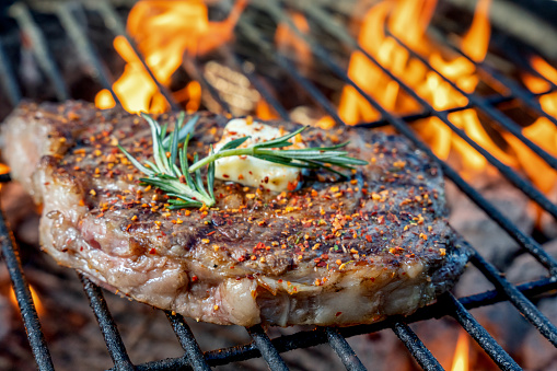 Grilled Ribeye Steak On A Hot Charcoal Grill With Flames Topped With Sauteed Garlic Cloves A Pat Of Butter And Rosemary