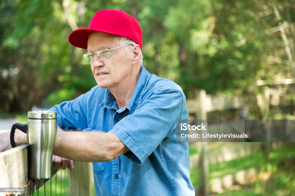 Caucasian senior man stands by wooden rail fence in rural scene. Caucasian senior man stands by wooden rail fence in rural scene.  He holds a reusable coffee mug.
He is relaxed and enjoying the morning quiet outside in the clean fresh air.  He wears a red baseball hat and a blue denim shirt.  He also wears glasses and has gray hair.  He is content. Baseball Cap Stock Photo