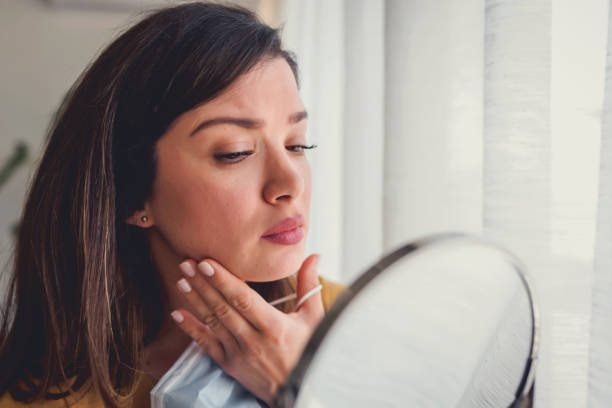 Woman looking herself in the mirror at home standing by the big window. She is concerned about acne, maskne Woman looking herself in the mirror at home standing by the big window. She is concerned about acne, maskne skin inflammation stock pictures, royalty-free photos & images