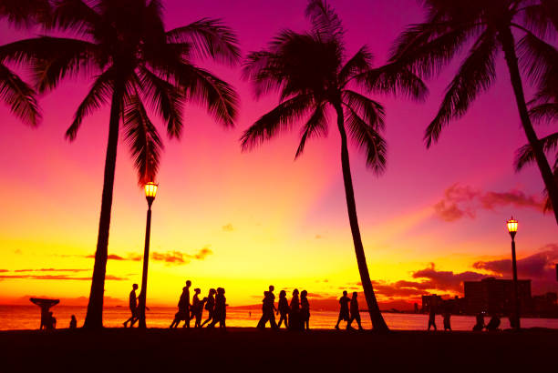 Sunset with vibrant colours at Waikiki Beach.\nPeople going home after a sunny day on the beach.\nSilhouettes of people and palm trees.