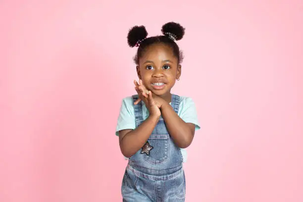 Happy laughing black skinned little girl in casual wear, jeans overalls, looking at camera, holding hands together. Little child with afrohair on pink studio background.