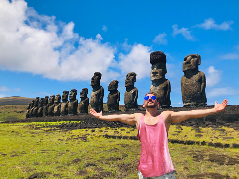 A handsome young happiness cheerful joyful fit backpacker latin male tourist visiting Ahu Tongariki Easter Island - Rapa Nui, in the middle of Pacific Ocean, part of Chile in Latin America; posing for a picture in front of the Moai's sculptures with his open outstretched arms at Ahu Tongariki, wearing a pink tank top t-shirt and blue stylish sunglasses, in a sunny summer hot day. Man visiting and sightseeing Latin America destination.\n\nLatin America / Chile travel concept.
