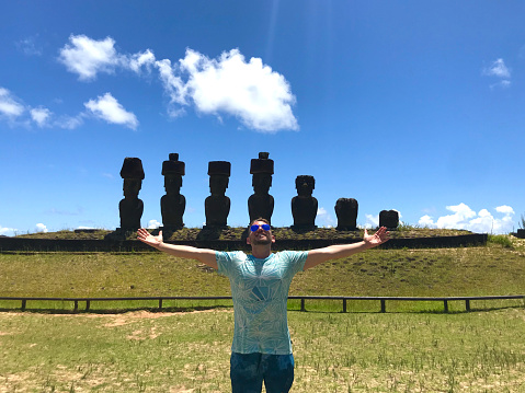 A handsome young happiness cheerful joyful fit backpacker latin male tourist visiting Anakena Beach, Easter Island - Rapa Nui, in the middle of Pacific Ocean, part of Chile in Latin America; posing for a picture in front of the Moai's sculptures with his open outstretched arms at Anakena Beach, wearing a blue t-shirt and blue stylish sunglasses, in a sunny summer hot day. Man visiting and sightseeing Latin America destination.\n\nLatin America / Chile travel concept.
