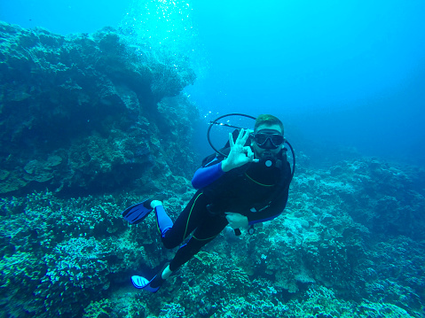 Adult Man with short brown hair wearing diving equipment underwater, holding a action camera while diving in the sea, seabed in the background