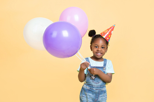 Portrait of little adorable happy black skinned kid girl with air balloons on bright yellow studio background. Birthday, holiday concept