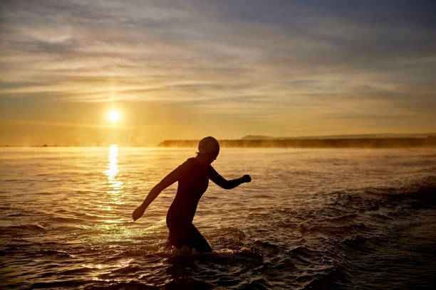 Partial view of early 50s Hispanic triathlete striding out of water after early morning workout with reflection of rising sun in background.