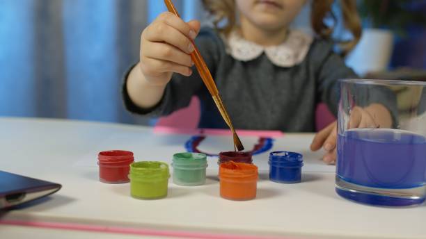 istock Child schoolgirl enthusiastically drawing with brushes, paints at home sitting at table using laptop 1303074622