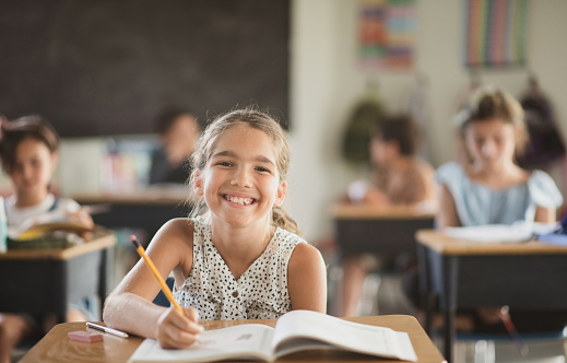 An elementary school girl is sitting at her desk in a classroom and smiling widely at the camera.