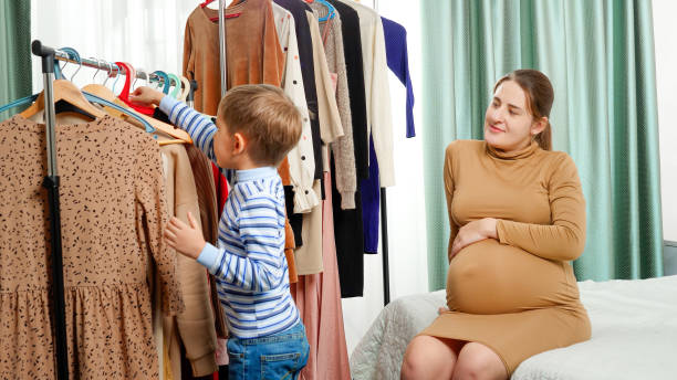 felice ragazzo sorridente prendendo vestiti da abiti rack pubblicità dandogli sua madre nel guardaroba - human pregnancy clothing shopping retail foto e immagini stock