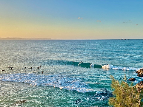 Horizontal high up aerial seascape of blue ocean waves and surfers with surfboards in green tropical paradise with pastel sunrise headland cliff trees in beautiful landscape of Clarke’s beach Byron Bay Australia